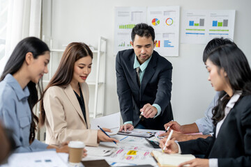 A group of people are sitting around a table with a man pointing at something