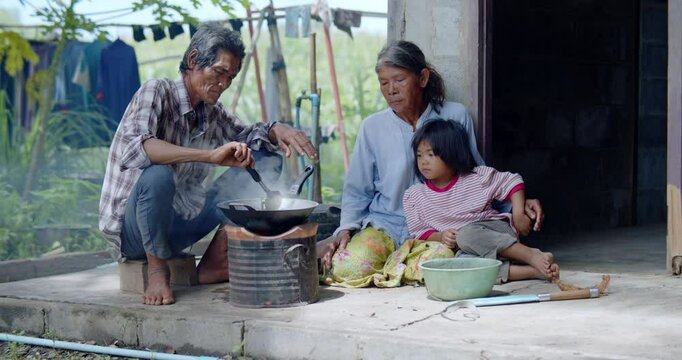 The simplicity of rural life of an Asian couple who are the poor farm laborers, cook dinner on a charcoal stove outside their rundown home in rural Thailand, while their granddaughter waits nearby. 
