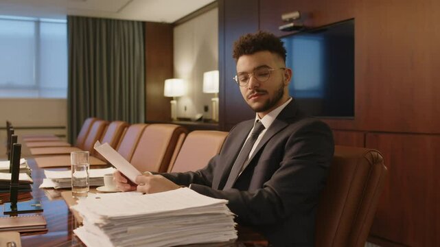 Portrait of young associate holding paper sheets looking at camera, sitting at wooden table with stack of documents in boardroom of law firm