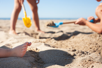 Close-up of toddler's foot standing on the beach sand and in the background children digging with toy shovels.