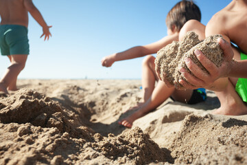 Close-up of a child's hands full of sand and his siblings playing in the background.