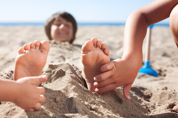 Children's hands are tickling the soles of the feet of a child who is buried in the beach sand.