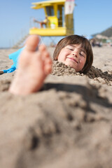 Portrait of a smiling child buried in the sand on the beach. Only his head and feet are sticking...