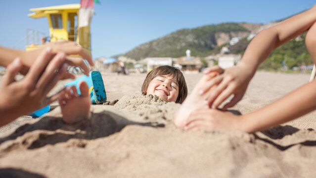Sand-covered boy smiles as hands tickle the soles of his feet.