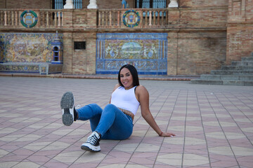 Young and pretty brunette Moroccan woman dressed in white top and jeans sitting on the ground with...