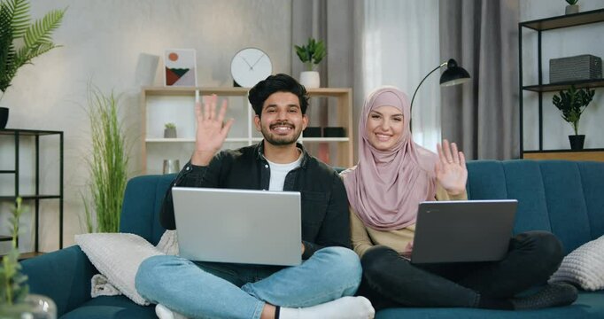 Multiethnic man and woman couple sitting in relaxed pose with computers on comfortable couch at home while looking into camera with waving hands