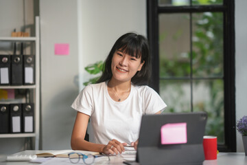 Fototapeta premium Asian businesswoman happily works on a tablet in a modern office, exuding confidence and cheerfulness as she tackles tasks surrounded by technology and paperwork, illuminated by natural light