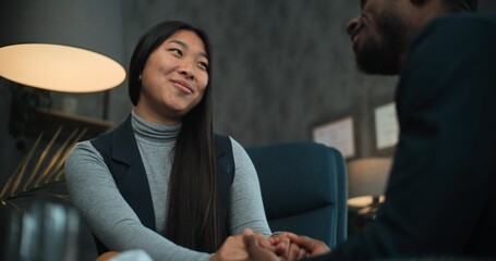 Asian woman smiles, talks to her boyfriend or husband during counseling session with therapist. Happy multiethnic couple sit on chairs holding hands, find compromise after quarrel on couples therapy.