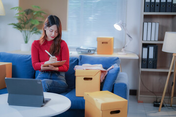 Woman working from home, managing her small business, checking inventory and writing on clipboard while sitting on a blue couch surrounded by cardboard boxes