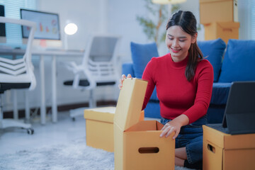 Asian woman happily opens a delivery box in her home office, showcasing her online store business. She works on her computer surrounded by items, exuding fulfillment and success in e-commerce