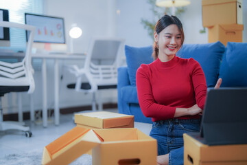 Asian woman happily unpacks boxes in new office, chatting on tablet during video call. Enjoying remote work in bright, modern space