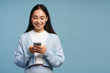 Smiling Asian woman holding mobile phone, using mobile app, standing isolated on blue background