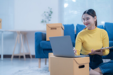 Young woman is using a laptop and holding a clipboard while sitting on the floor of her new apartment, surrounded by moving boxes
