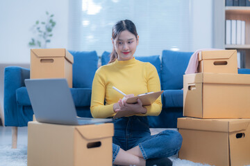 Young woman is sitting on the floor and managing her online business from home. She is writing on a clipboard and using a laptop