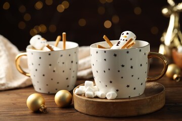 Cute marshmallow snowmen in cups of hot drinks on wooden table with Christmas decor against blurred lights, closeup
