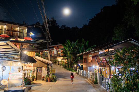 A woman walks down a street at night in Mae Kampong village with a full moon in the sky, Thailand