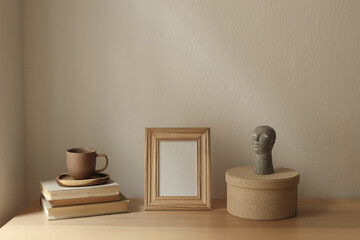 Artistic boho interior still life. Blank wooden photo frame mockup on table. Scandi interior decor. Cup of tea, coffee, old books. Boho busta sculpture, statue. Round gift box. Beige wall background