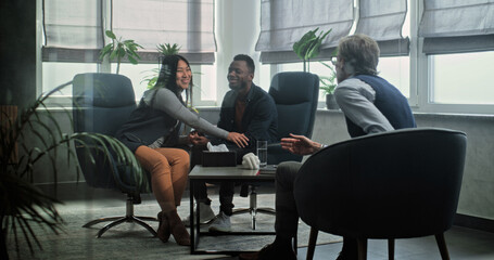 Multiethnic couple on counseling session with therapist. Mature psychologist talks to young family. African American man and Asian woman sit on chairs holding hands after quarrel on couples therapy.
