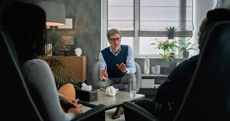 Mature psychotherapist talks to young family, provides couples therapy. Back view of multiethnic couple sitting on chairs, listening psychologist on counseling session. Psychology and sexology concept