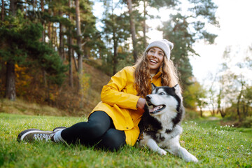 A young woman wearing a yellow jacket and a knit hat smiles as she plays with her Siberian husky in a vibrant forest filled with autumn colors. Concept of fun, entertainment.