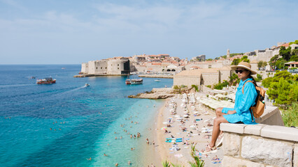 Obraz premium A traveler enjoys the serene beauty of Dubrovnik's vibrant shoreline, surrounded by crystal-clear waters and bustling activity. Sunbathers relish the picturesque beach beneath a radiant blue sky.