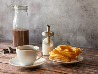 Still life of a cup of hot chocolate with churros, glass bottle with chocolate at the bottom and glass sugar bowl. Wooden table