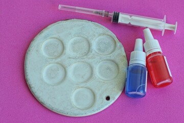 one round old iron medical white laboratory Petri dish with cells for research of biological material with two plastic small plastic bottles with blue and red liquid and syringe lie on pink table