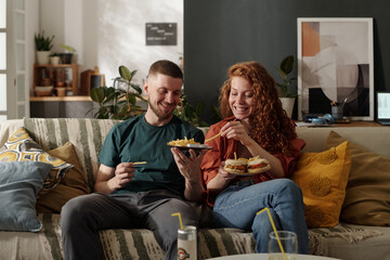 Smiling young man sharing french fries with his pretty girlfriend holding plate with hamburgers...