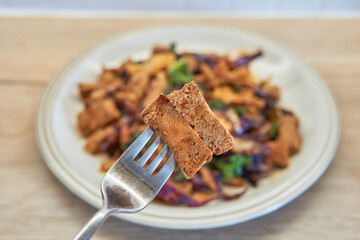A close-up shot of a fork holding a piece of tofu above a plate of colorful stir-fried vegetables, showcasing a delicious vegan meal.
