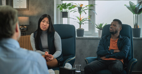 Obraz premium Asian young woman talks about relationship conflict. Multiethnic family on counseling session with psychologist. In the foreground, unfocused therapist talks to young couple during couples therapy