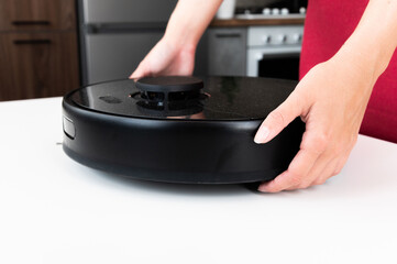 Black robot vacuum cleaner in female hands, close-up. The woman puts the vacuum cleaner on the table to disassemble and clean it after cleaning