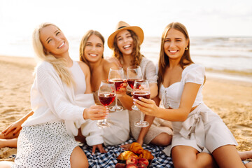 Four friends celebrate together at the beach, raising their glasses of red wine. The sun sets in the background, creating a warm and joyful atmosphere. Hen party.