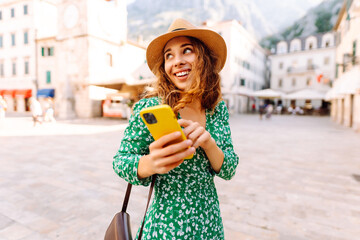 Young woman in a green dress smiles while using her yellow smartphone in a quaint European plaza on...
