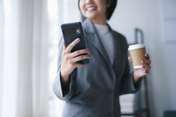 Smiling businesswoman in her office, holding coffee and using a smartphone. She takes a break to browse the internet, creating a modern professional lifestyle vibe