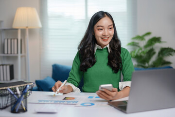 Asian businesswoman is working remotely, analyzing graphs and using her smartphone at her home...