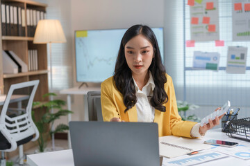 Asian businesswoman is sitting at her desk using a laptop and holding a calculator while working on finances