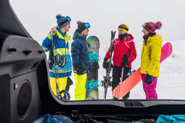 Fototapeta premium Group of friends with snowboards and skis chatting near car trunk
