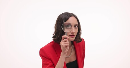Curious woman in red blazer looks through magnifying glass on white background in studio. Brunette female playfully demonstrates attention to details - Powered by Adobe