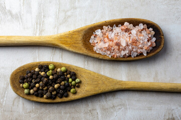 A pair of wooden spoons on a neutral tile background.  Rustic spoons are filled with Himalayan salt and peppercorns.
