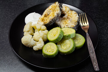Spiced cod fish steaks, cauliflower and zucchini steamed with sour cream sauce on black ceramic plate with gilded fork close-up view from top. A healthy meal for lunch.