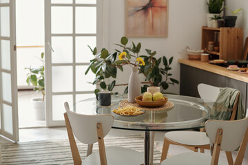 Round glass table with french fries, vase with flowers, two mugs and bowl with fresh fruits standing in the center of cozy kitchen