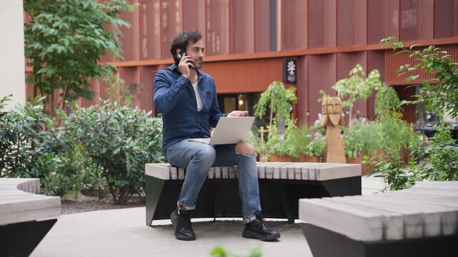 Attractive Caucasian male spending free time in middle of urban square. Talking on smartphone with colleague or friend while using laptop on his knees. Spending free time with joy. Sunny weather.