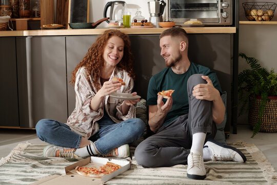 Happy young couple sitting on the floor by kitchen counter and enjoying appetizing takeout pizza during brunch or lunch - Powered by Adobe