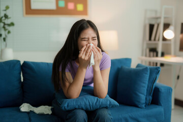 Young woman is suffering from a cold and blowing her nose with a tissue while sitting on her sofa