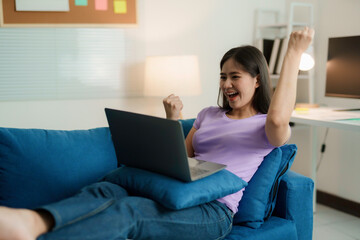 Naklejka premium Young asian freelancer is raising her arms in celebration while working on her laptop, expressing joy and excitement in her home office