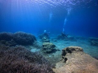 Obraz premium Rocky underwater landscape with pair of swimming scuba divers. Underwater photography from scuba diving exploration trip. Divers in the ocean, travel photo. Bubbles from breathing in the water.
