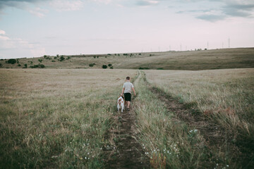 A boy of about seven with blond hair moves through a meadow with an Epagnol Breton dog against a background of grass and hills at sunset. Rear view. 
