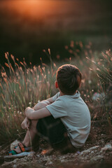 A boy in nature, about seven years old, rear view, with blond hair, wearing a beige T-shirt and black shorts, sitting on the grass against the background of the sunset. 