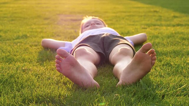 The child is lying on the green grass. The child is swinging their bare feet. The child is enjoying nature and relaxing outdoors. The concept is a happy childhood and a family picnic.