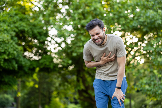 Young Indian man doing sports and jogging in the park, looking forward, holding hand to chest and grimacing from severe heart pain